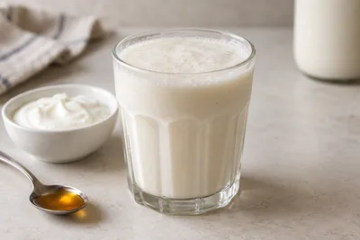 Milk-shake au yaourt blanc dans un verre cannelé, avec un bol de yaourt et une cuillère de miel.
