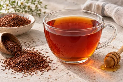 Théière en verre remplie de rooibos infusé, couleur rouge cuivré, avec une gousse de vanille posée à côté